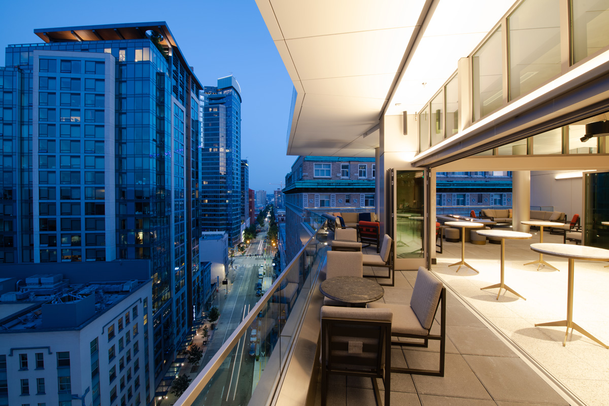 A balcony with a glass railing and seating area overlooking a city street at dusk.